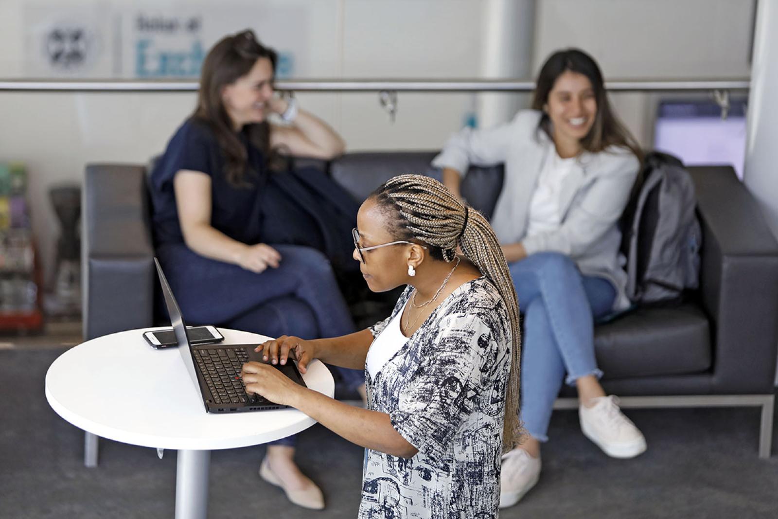 Student working at a round desk on laptop with two students socialising on a sofa in the background