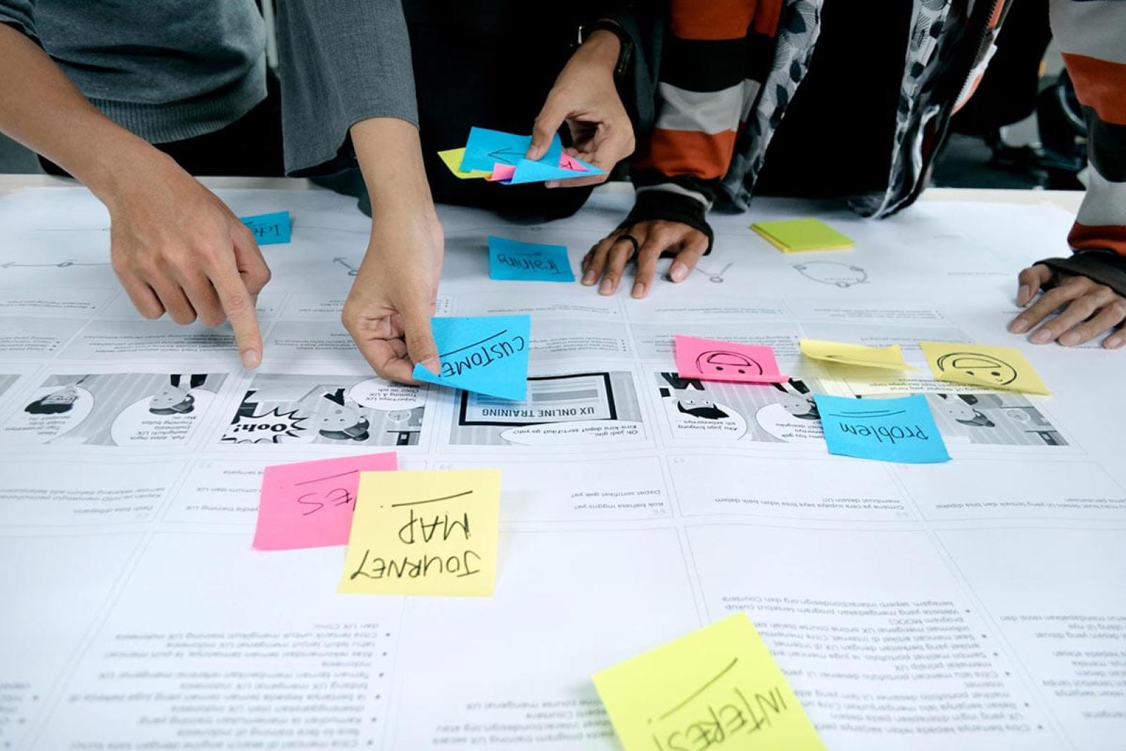 Arms of people standing over a desk covered with paperwork and coloured sticky notes